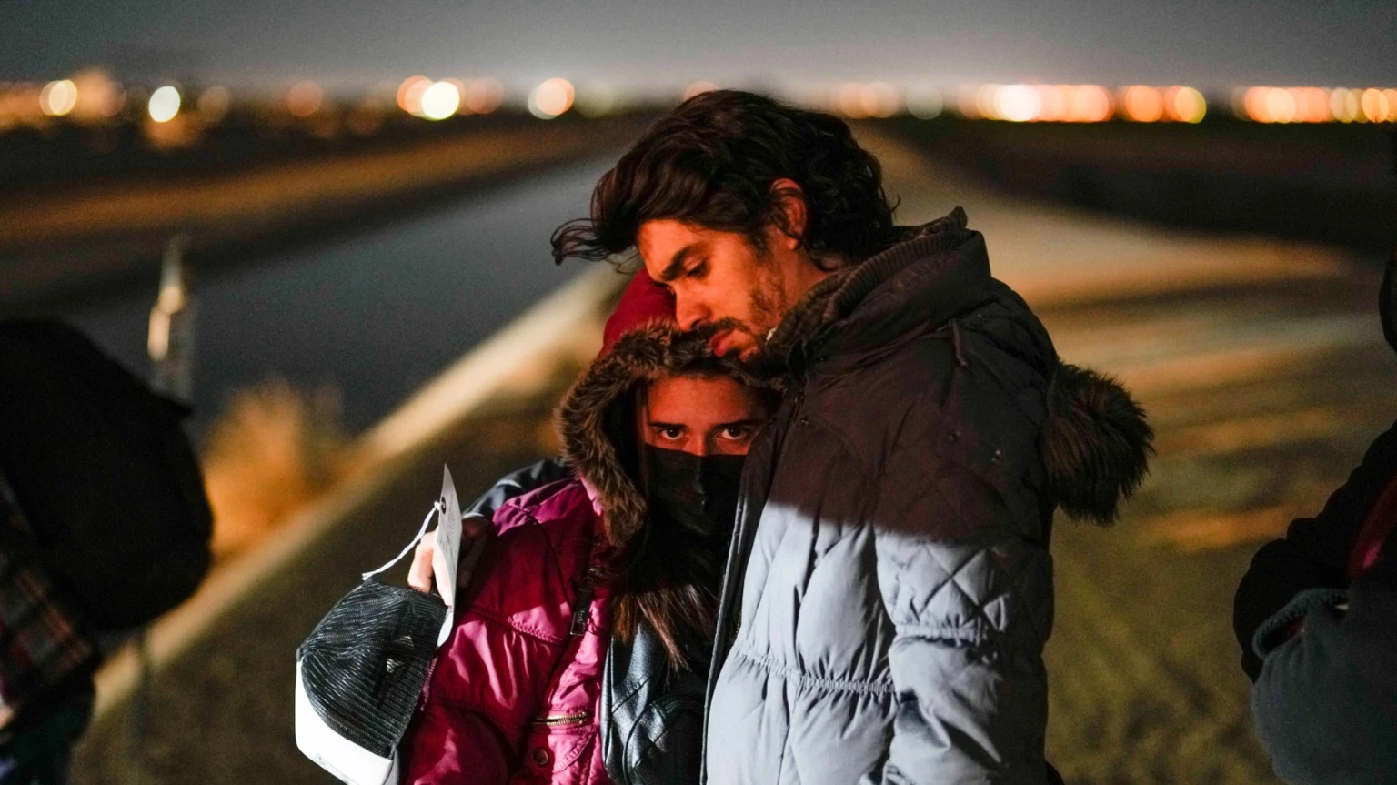 A woman looks at the camera as she leans on a man who has his arm around her. The couple wait to be processed for asylum after crossing the border into the U.S.